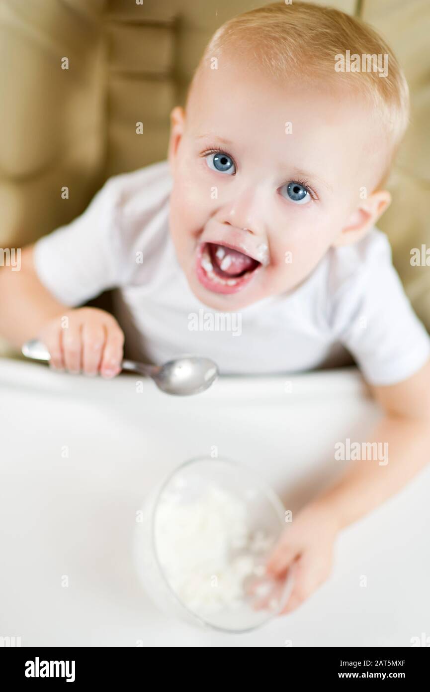 Ein kleines blauäugiges Mädchen in einem Fütterstuhl legt einen Löffel mit Reisporridge in den Mund. Ansicht von oben, vertikales Foto Stockfoto