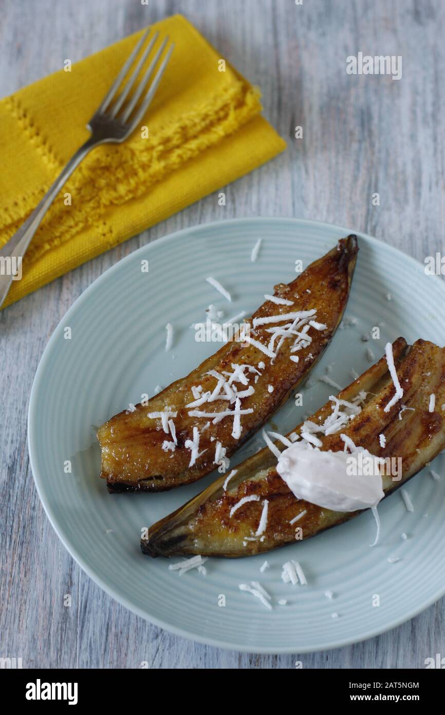 Goldbraun gebackene Banane halbiert mit geriebener Kokosnuss und Joghurt auf blauem Teller mit gelber Serviette und Gabel Stockfoto