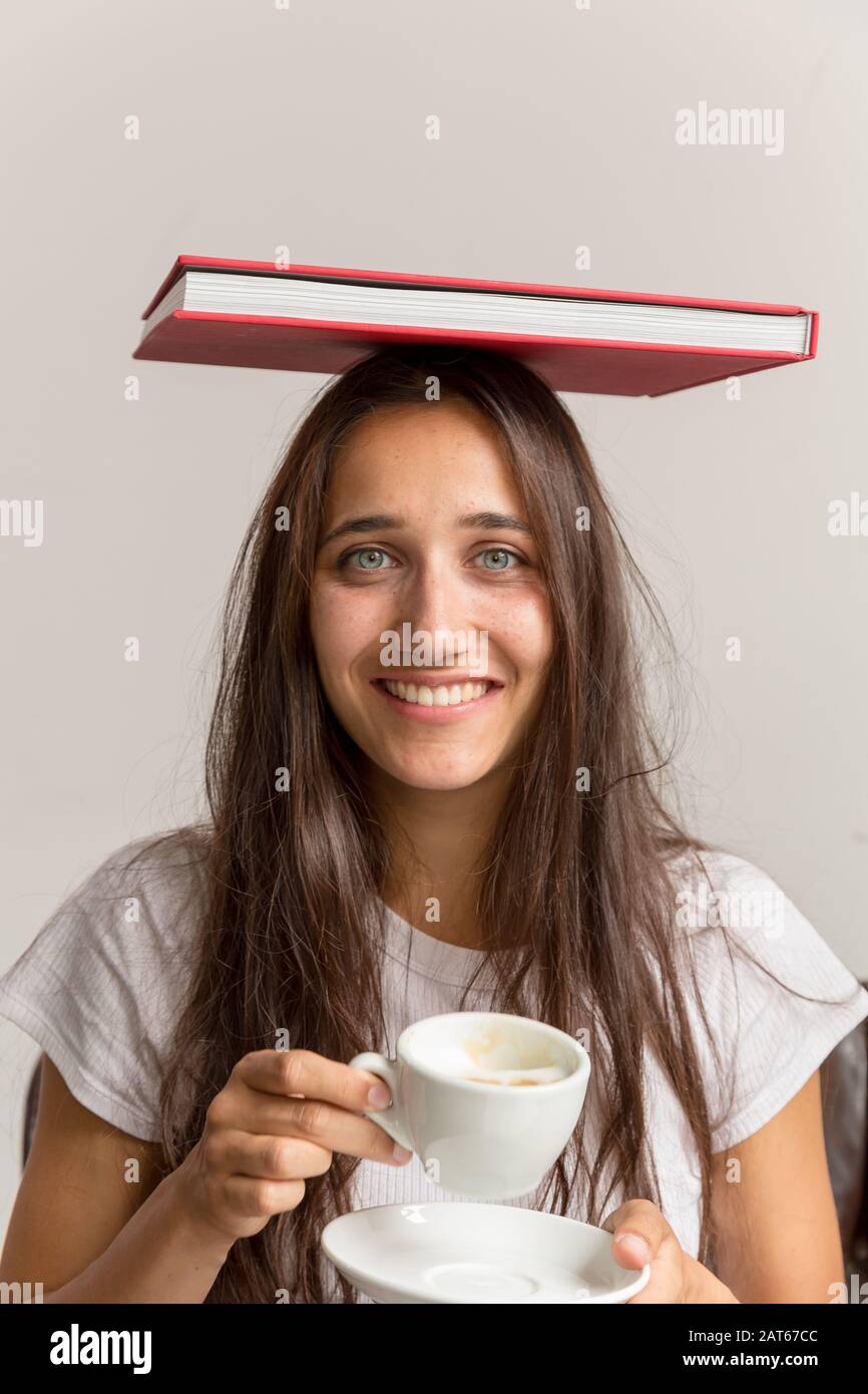 Junge Frau mit gemischter Rasse indische Ethnie, die Buch auf dem Kopf balanciert und die Kaffeetasse weithin lächelnd hält. Mittlere Aufnahme. Stockfoto