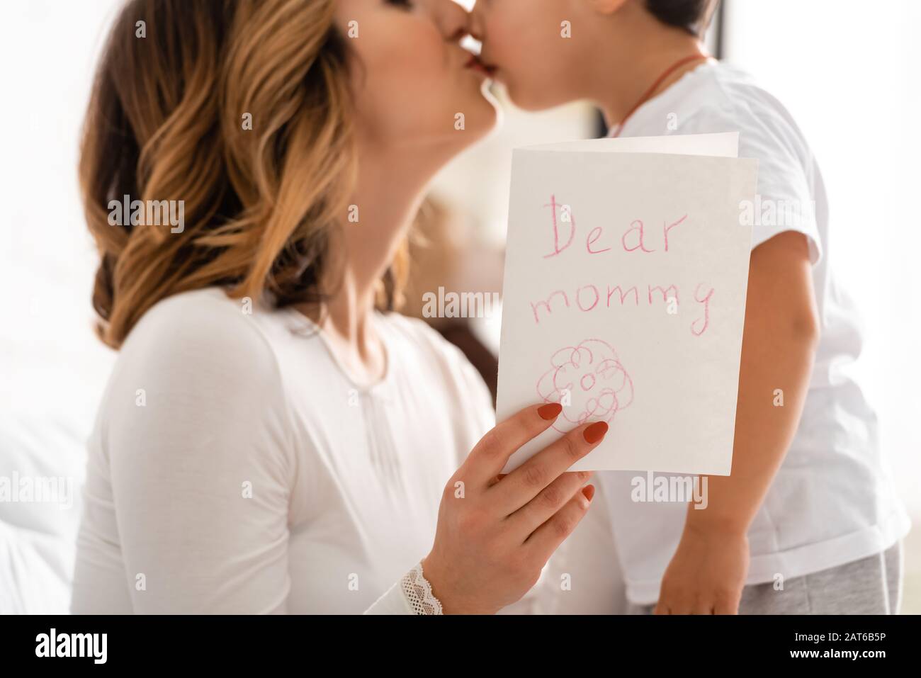 Beschnittener Blick auf die glückliche Mutter, die den liebenswürdigen Sohn küsst, während sie die Tageskarte der Mütter mit der Aufschrift "Liebe Mama" hält Stockfoto