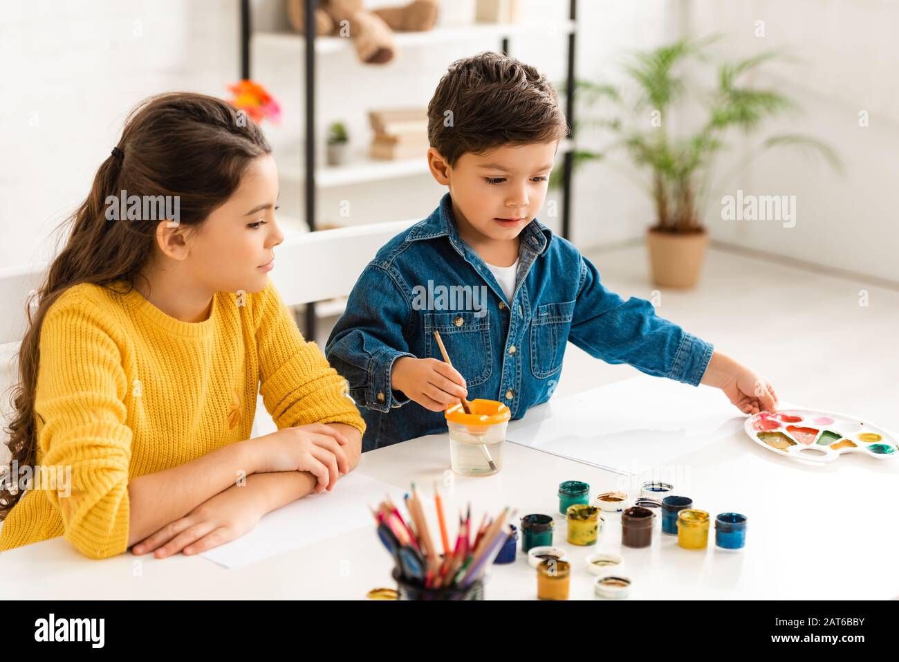 Süßer Junge, der Pinsel in Wasser taucht und die Palette berührt, während er am Tisch in der Nähe der Schwester sitzt Stockfoto