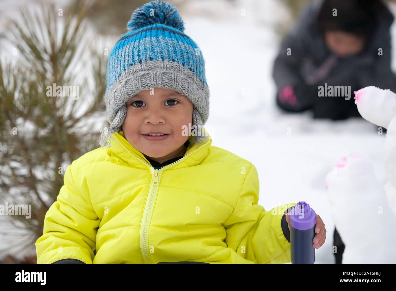 Ein kleiner Junge mit einem niedlichen Lächeln, das sich in einem schneebedeckten Berg befindet, farbenfrohe Winterkleidung trägt und einen Schneemann mit Farben malt. Stockfoto