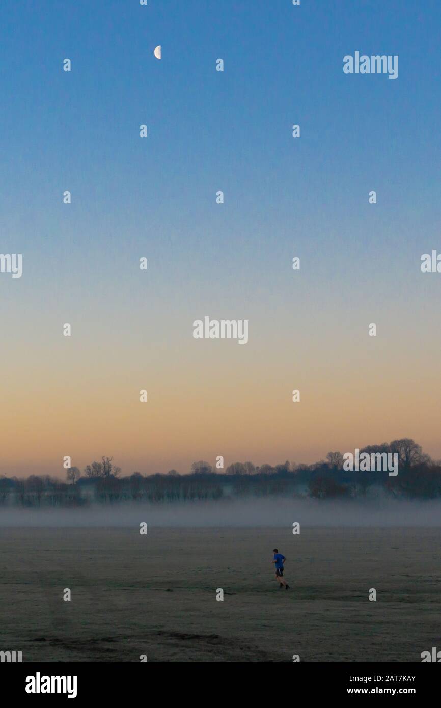 Person, die bei Sonnenaufgang über ein nebeliges Feld läuft, wobei der Mond zu sehen ist Stockfoto