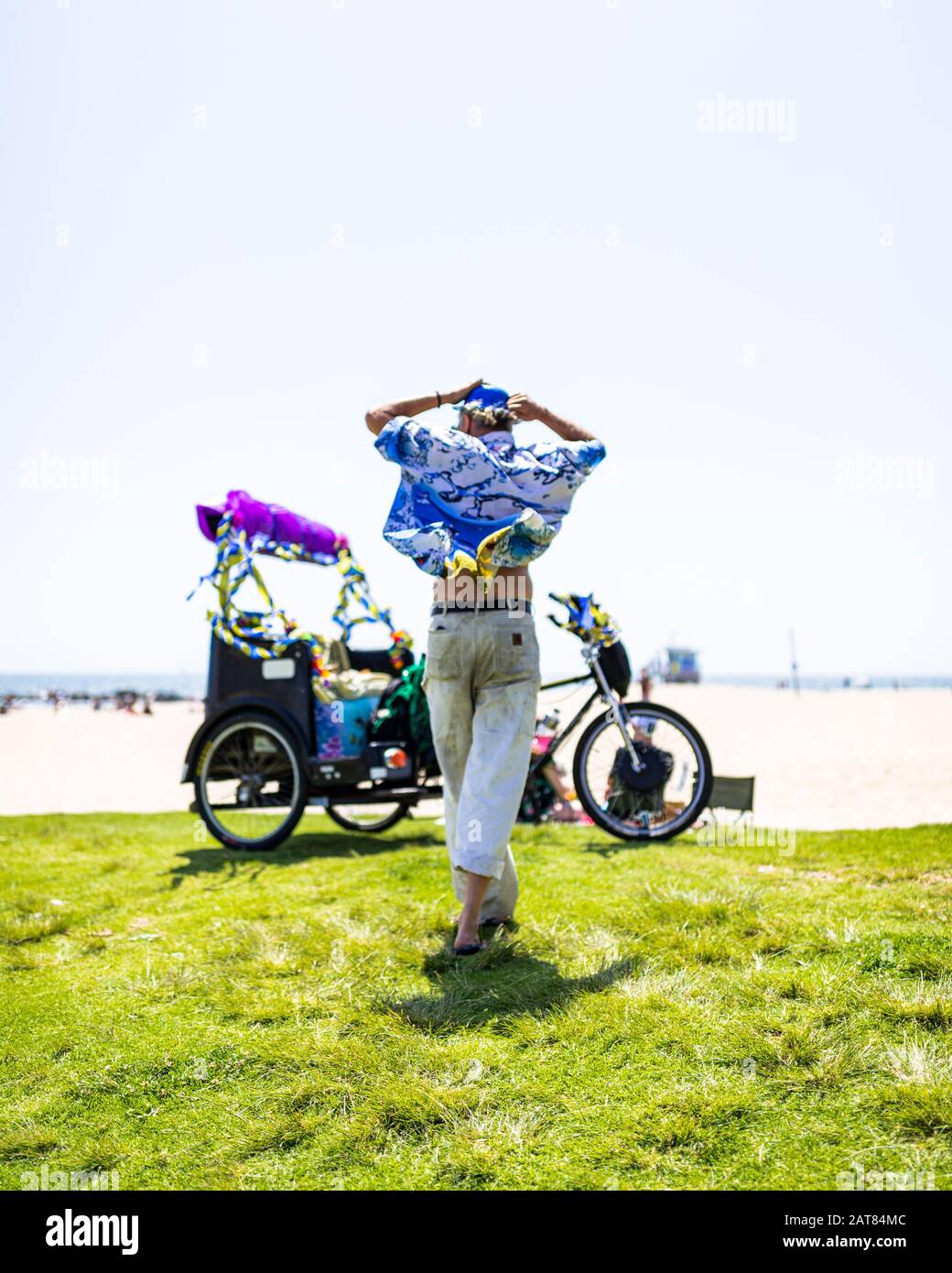 Einsamer Mann in Lebhaft Gefärbter und Festlicher Kleidung, Der Auf Gras Zu Einem Dekorierten Fahrrad am Strand von Venedig Mit Blick auf den Pazifischen Ozean läuft. Stockfoto