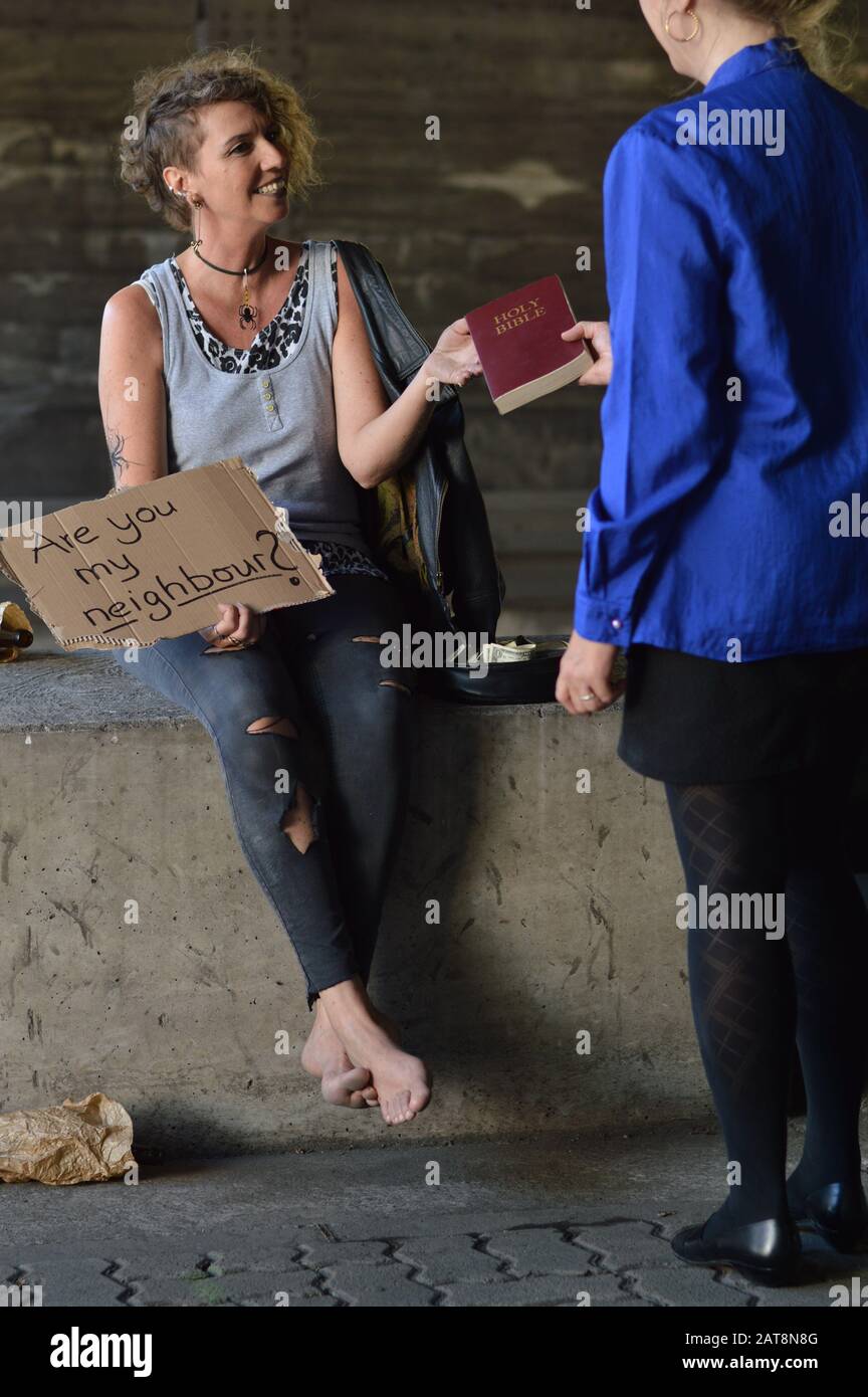 Eine Punk-Frau, die neben einer Flasche Alkohol unter einer Brücke sitzt, indem sie ein Schild hält und sich über eine bibel von einer Geschäftsfrau freut Stockfoto