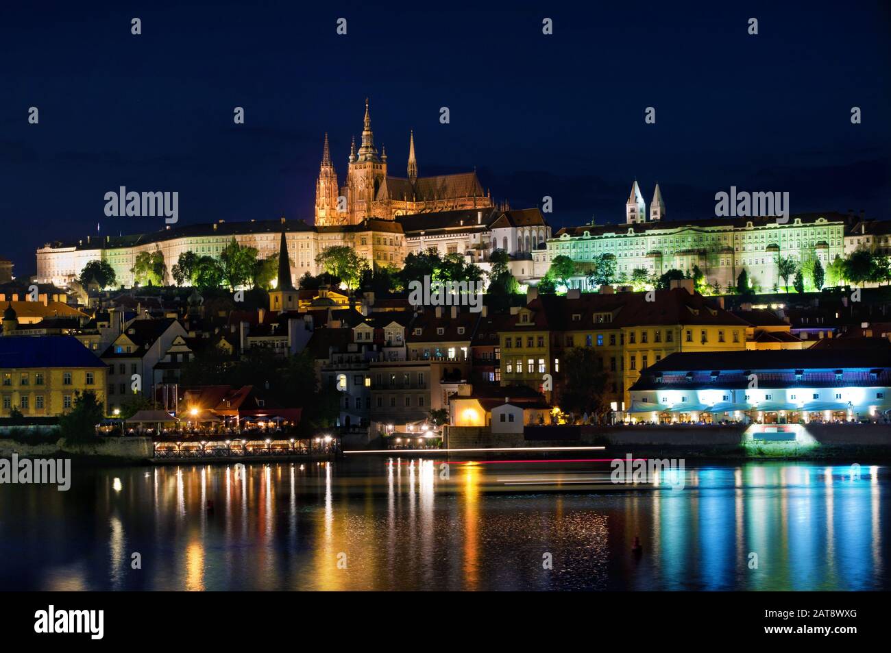 Prager Burg und Veitsdom mit vielen kleinen Häusern in der Nähe der Moldau gegen den dunkelblauen Nachthimmel, Spiegelung der Lichter im Wasser. Prag, Stockfoto