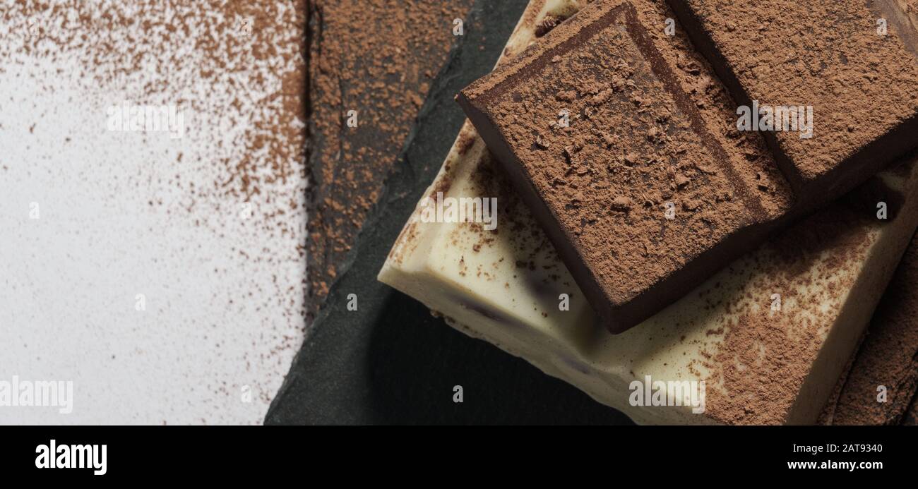 Dunkle und weiße Schokoriegel auf dunklen Steinplatten, die mit Pulverschokolade und Kakao bedeckt sind Stockfoto