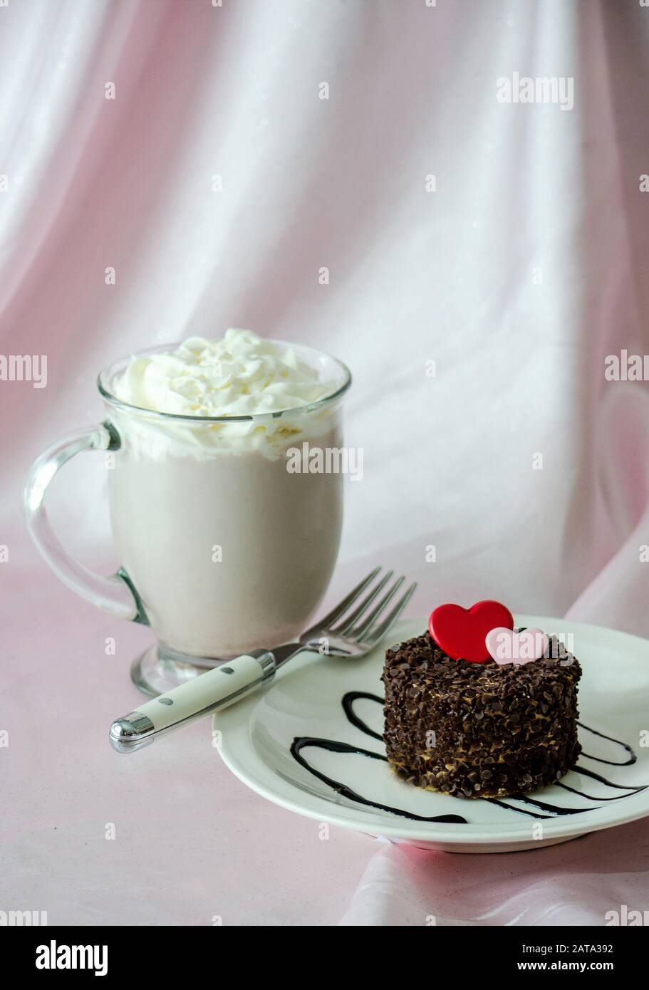 Ein Kuchen mit winzigen Schokoladenchips und einem schokoladenroten und rosafarbenen Herzen. Und ein Becher heißer Schokolade mit Schlagsahne. Süße Leckereien! Stockfoto