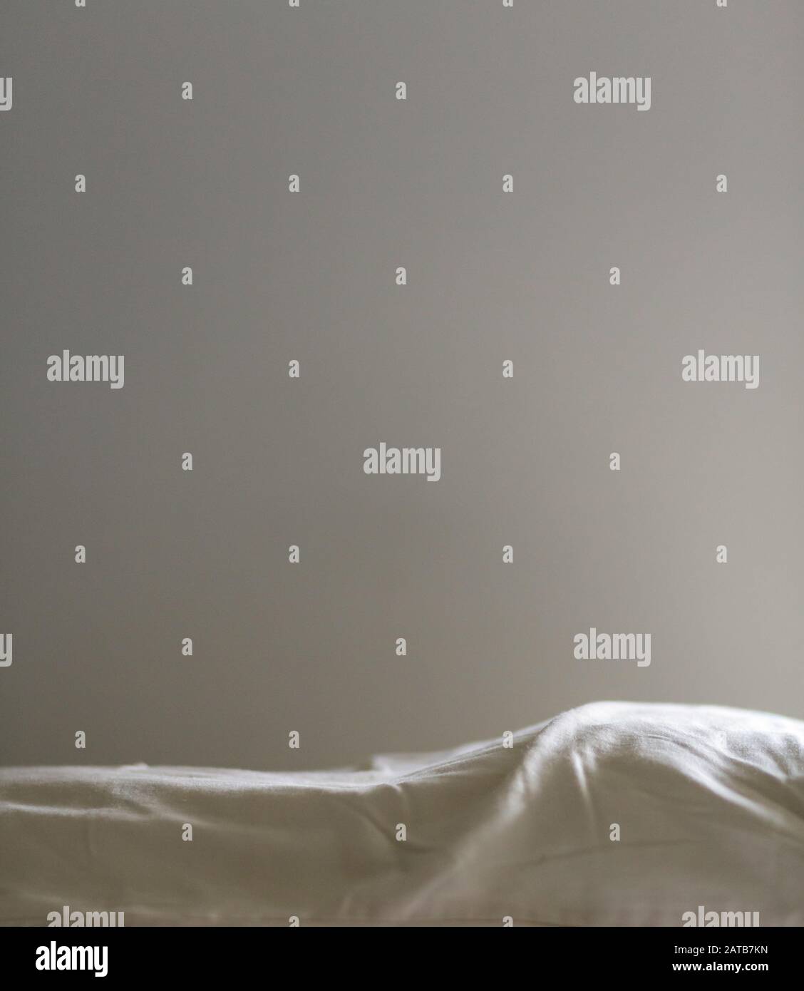 Eine weiche, helle monochromatische Wand aus gedämpftem Weiß über einem Hügel weicher Bettlaken in Off-Weiß, in Seitenlicht aus einem Morgenfenster zu sehen. Stockfoto