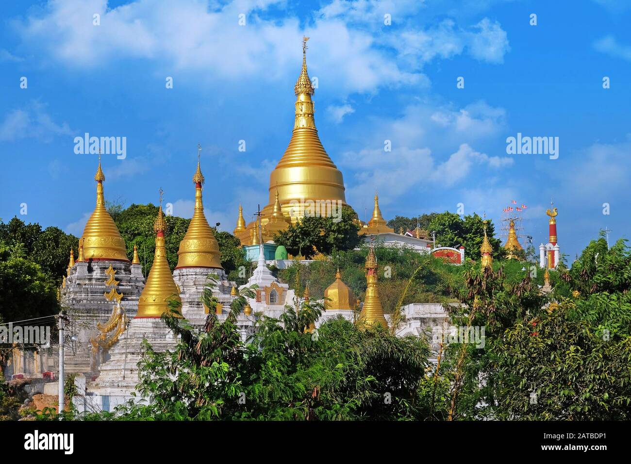Blick auf das goldene Dach einer birmanischen Pagode in Mingun, Mandalay, Myanmar, umgeben von grüner Vegetation, gegen einen blauen Himmel, der von weißen Wolken bedeckt ist. Stockfoto