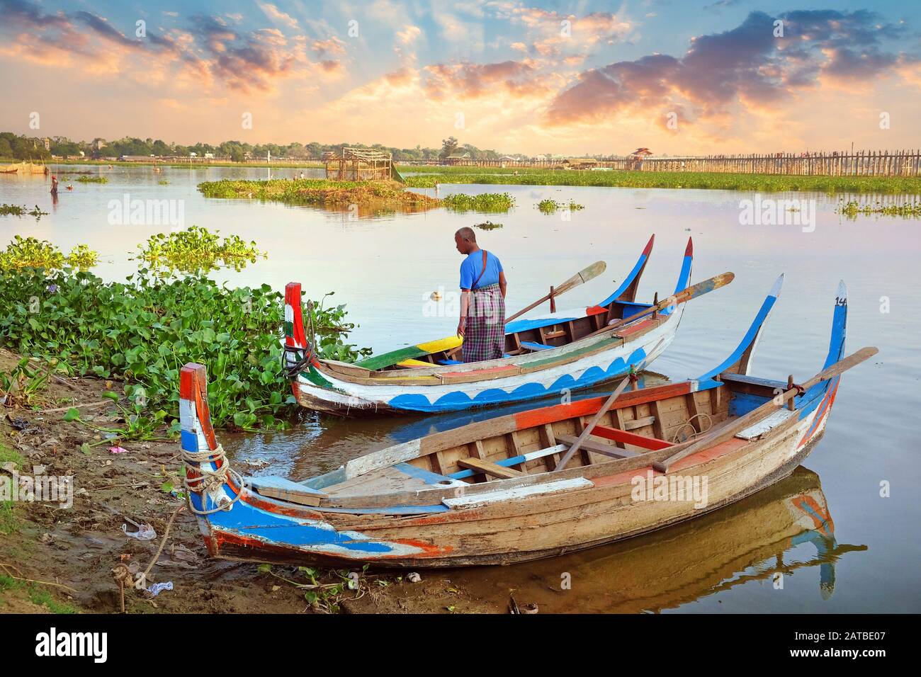Bunte traditionelle Boote auf der Seite des Taung Tha Man Lake, in der Nähe von Mandalay, Myanmar, umgeben von grüner Vegetation, gegen eine schöne bunte Stockfoto