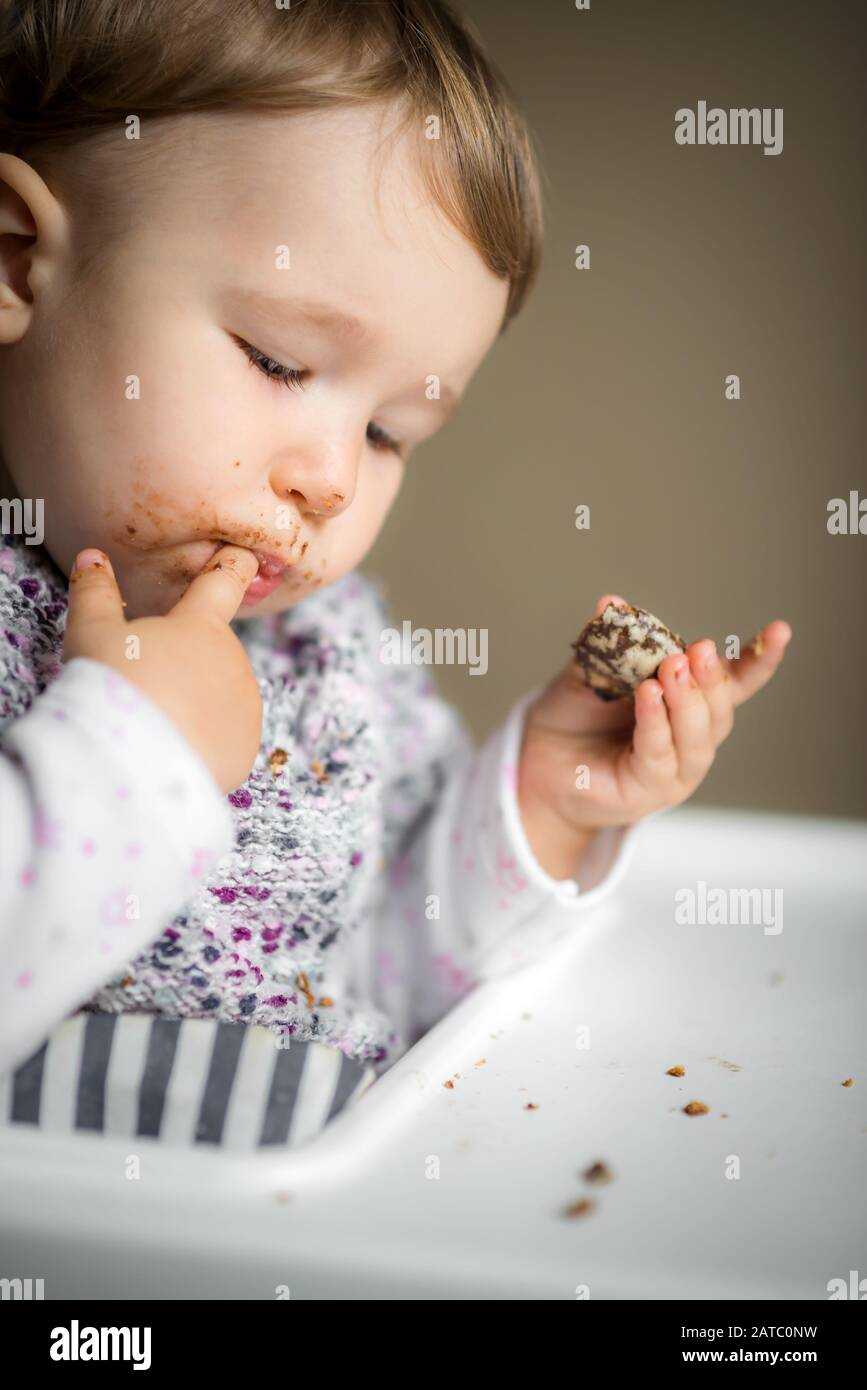 Baby Mädchen mit unordentlichen Gesichtern essen. Das einjährige Kind isst Kuchen und steckt sich die Finger in den Mund. Stockfoto