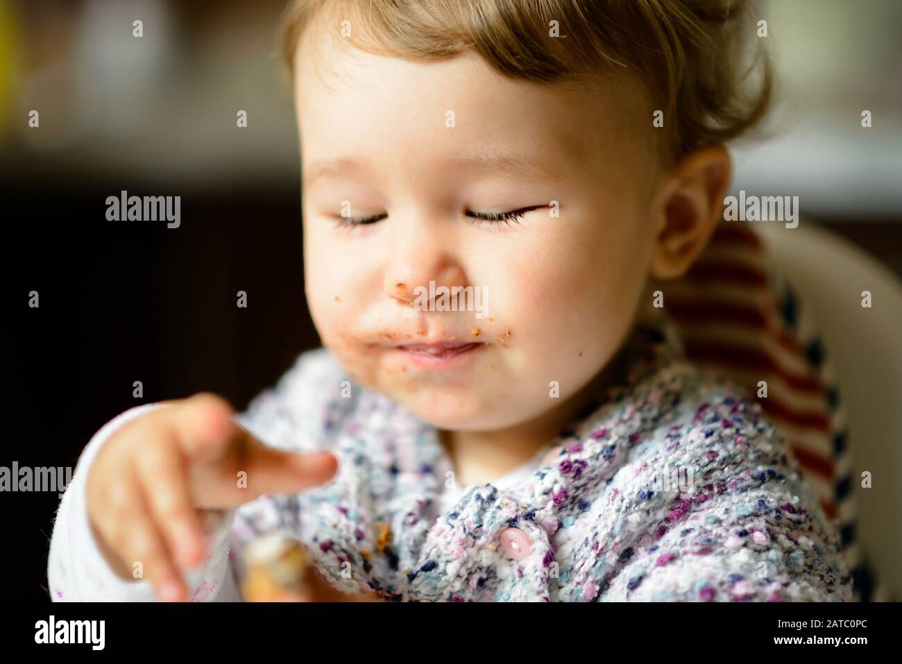 Fröhliches Baby-Mädchen mit unordlichem Gesicht essen. Das einjährige Kind isst und lächelt mit geschlossenen Augen. Stockfoto