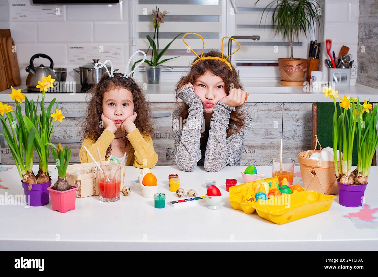 Zwei Schwestern malen Eier in der Küche. Frohe Familie, die sich auf Ostern vorbereitet. Niedliche Mädchen tragen bunte Ohren Stockfoto