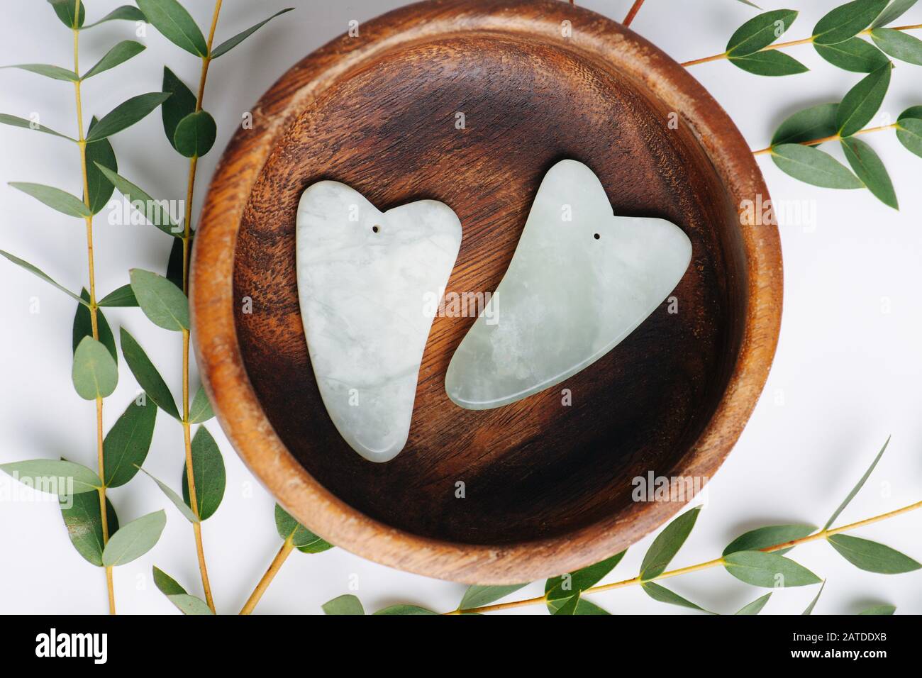 Draufsicht über Gua sha-Jadesteine in Holzschale über Ästen auf Weiß. Stockfoto