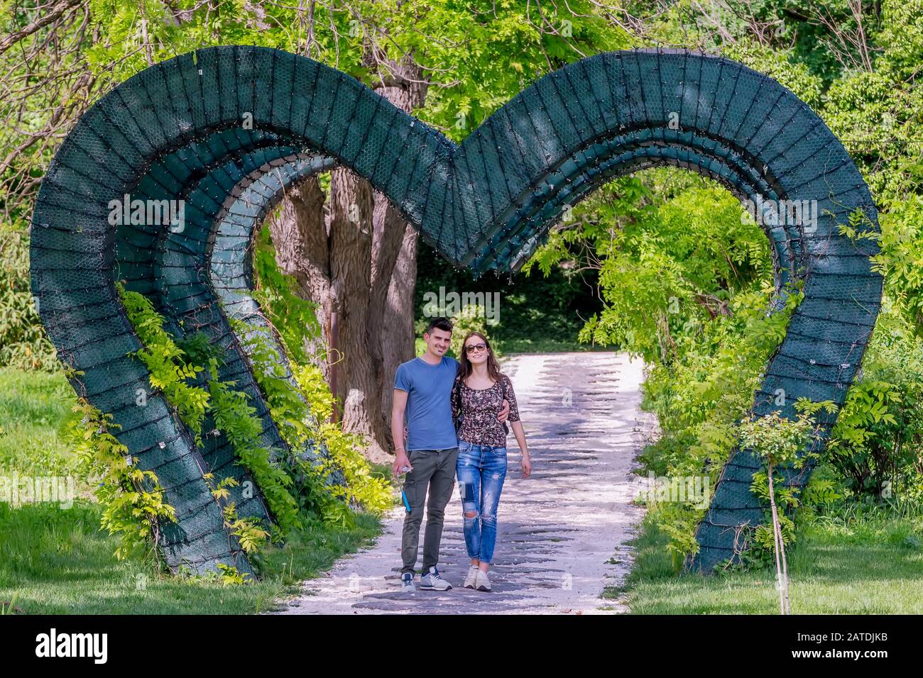 Das junge, engagierte Paar spazieren unter einem grünen, herzförmigen Bogen im öffentlichen Park Craiova, Rumänien Stockfoto