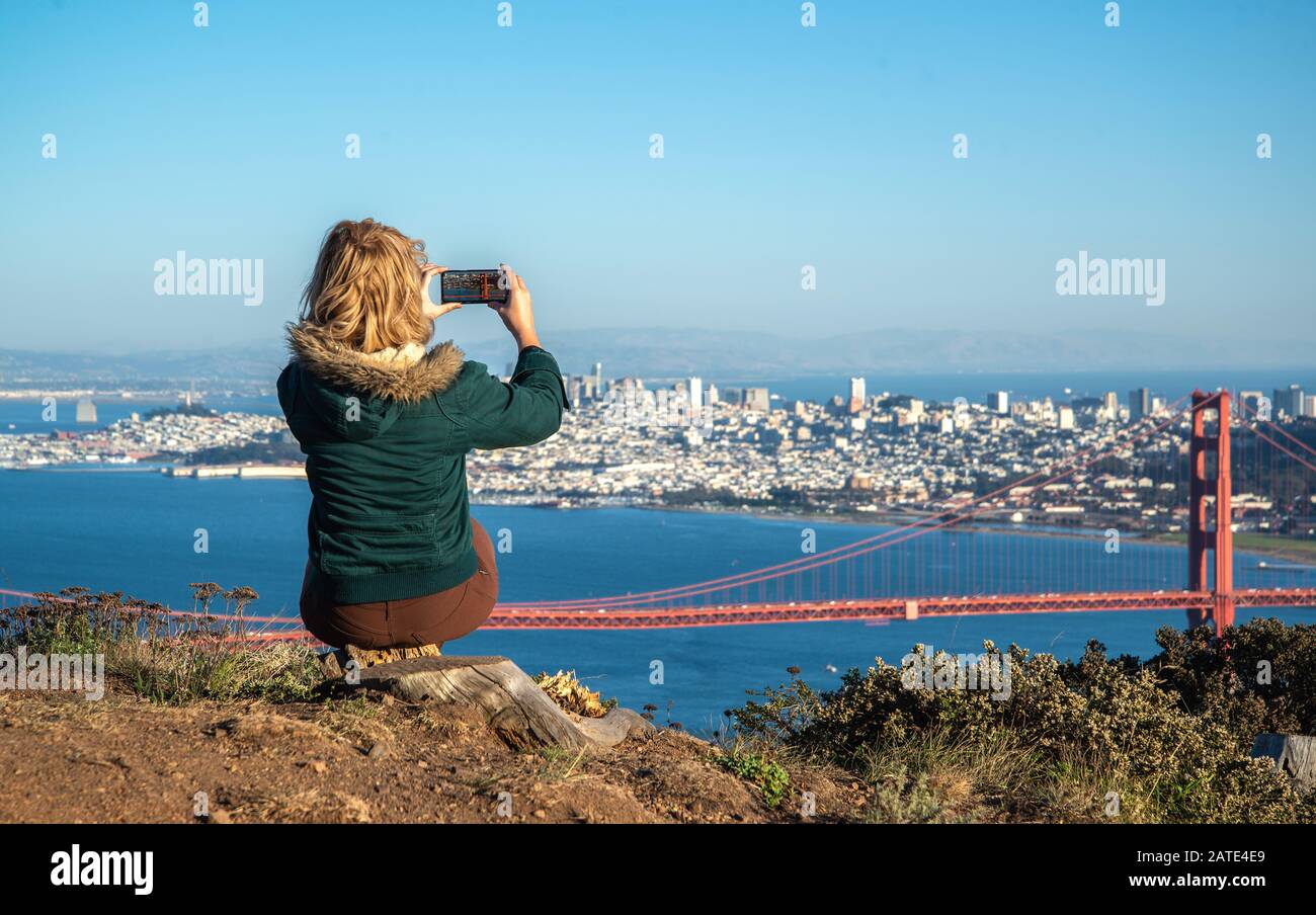 Junge blonde Frau in grüner Jacke, die mit ihrem Telefon der Golden Gate Bridge in San Francisco, Kalifornien, ein Foto macht. Tourist Foto von Golden Ga Stockfoto