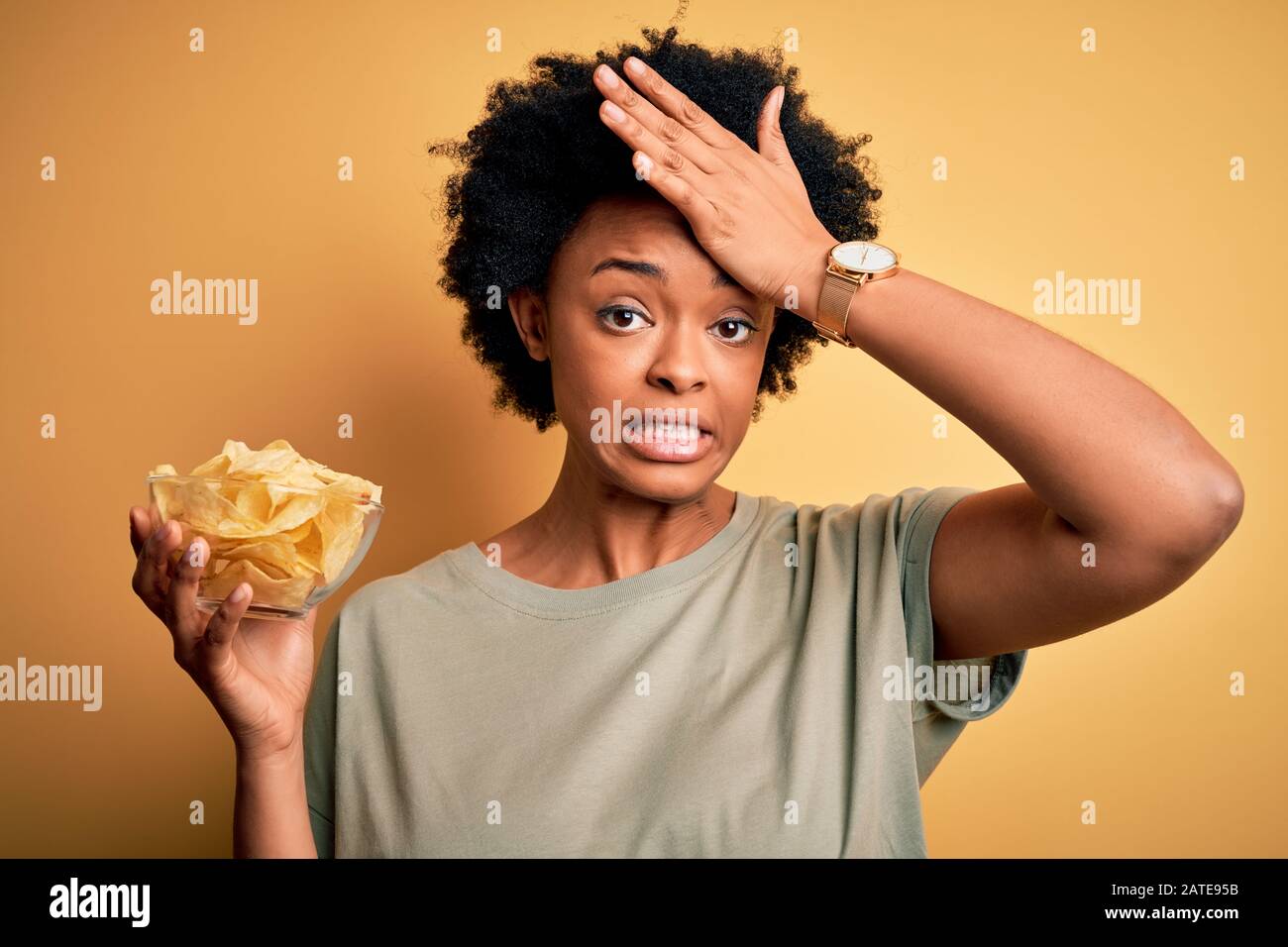 Junge afro-amerikanische afro-amerikanische afro-Frau mit lockiger Haarhalteschale mit Pommes Kartoffeln, die mit der Hand auf dem Kopf gestresst sind, schockiert mit Scham und Überraschungsgesicht, a Stockfoto