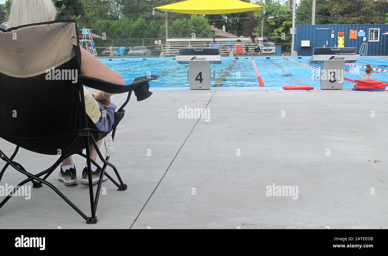 Frau mit Hut sitzt auf einem schwarzen Stuhl und beobachtet Schwimmer im Pool Stockfoto