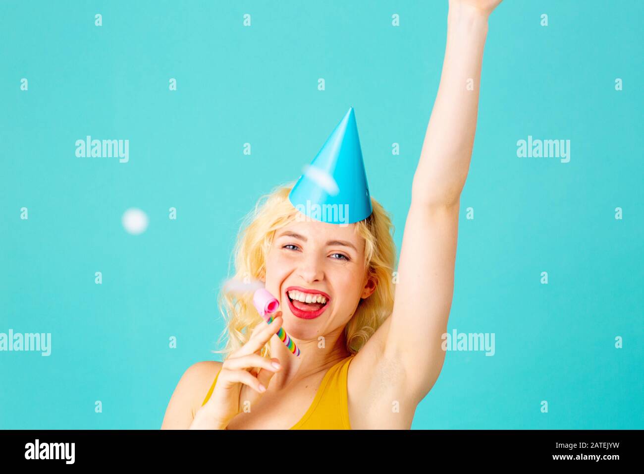 Porträt einer fröhlichen, lächelnden jungen Frau, die einen schönen Geburtstag feiert, mit Hut und Arm oben, isoliert auf Blau Stockfoto