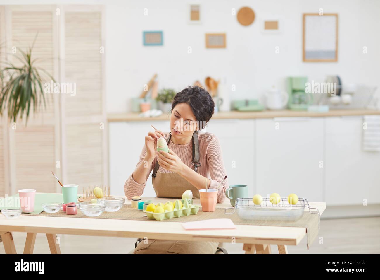 Weitwinkelansicht bei eleganter, erwachsener Frau, die Ostern Eier in Pastellfarben malt, während sie in Küche oder Kunststudio am Tisch sitzt, Kopierer Stockfoto