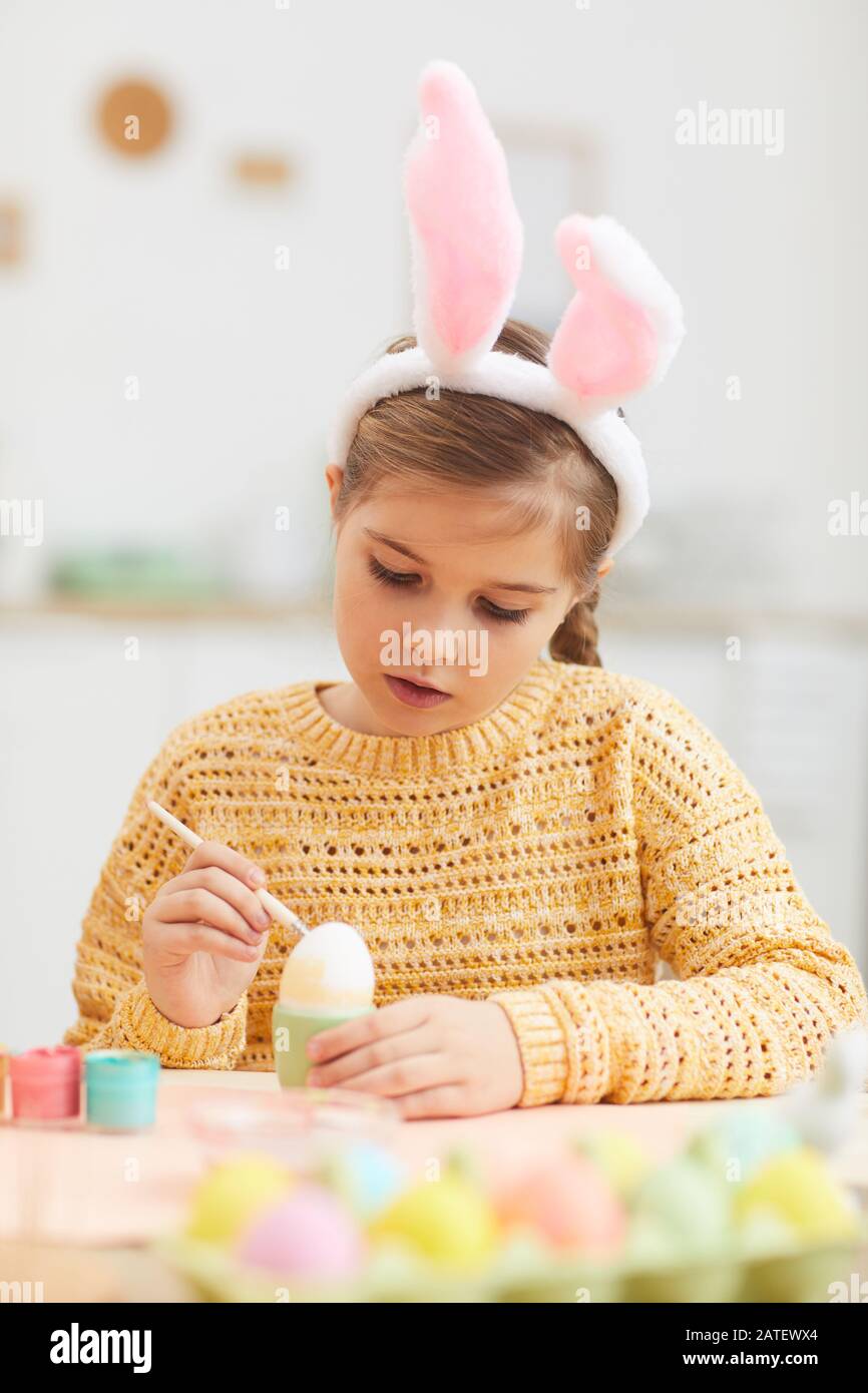 Vertikales Porträt des süßen kleinen Mädchens, das Ostereier malt, während er in der gemütlichen Kücheneinrichtung bunte Ohren trägt, Kopierer Stockfoto
