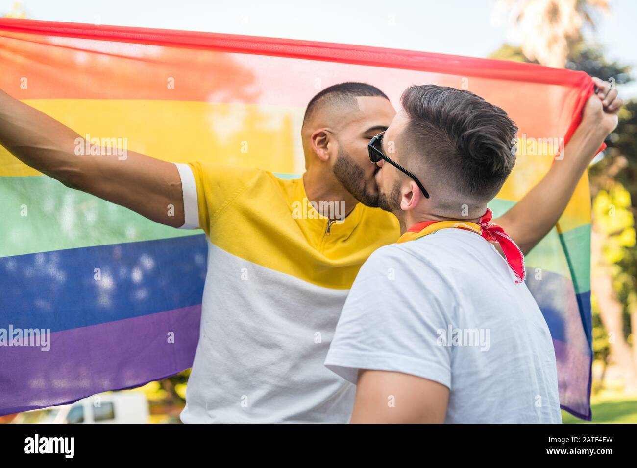 Porträt des jungen schwulen Paares, das sich küsst und seine Liebe zur Regenbogenfahne im Stret zeigt. LGBT und Love Konzept. Stockfoto