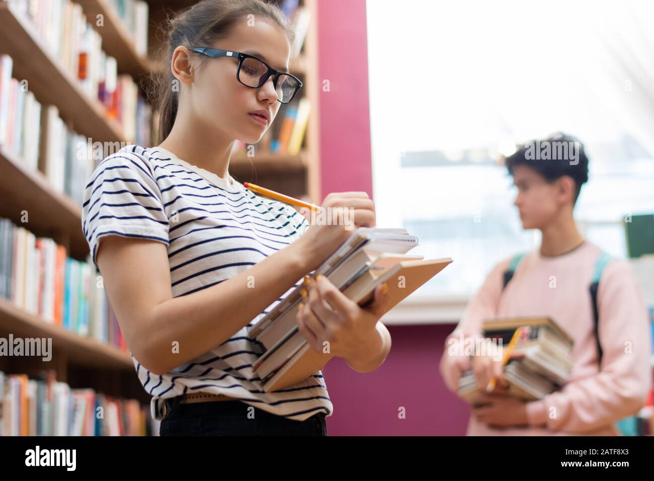 Cleverer Teenager in Brillen, der Notizen in Notizblock auf dem Stapel von Büchern macht Stockfoto