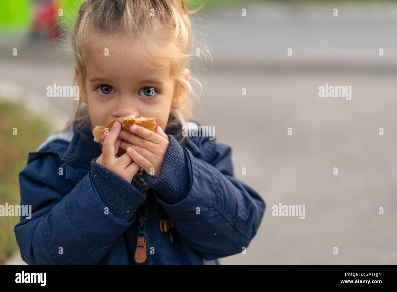 Ein wunderschönes kleines kaukasisches Mädchen mit blonden Haaren und dem Essen von Brot, das eifrig mit den Händen ist, blickt mit traurigen Augen, einem verlassenen Kind und hu auf die Kamera Stockfoto