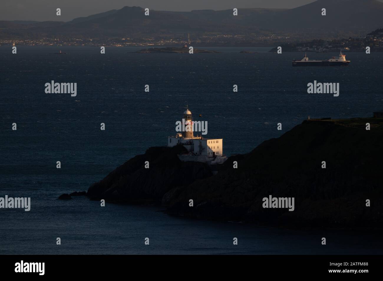 Der Baily Leuchtturm, Howth. co. Dublin, Baily Leuchtturm auf Howth Klippen, Blick auf den Baily Leuchtturm von der Klippe mit Frachtschiff im Hintergrund Stockfoto