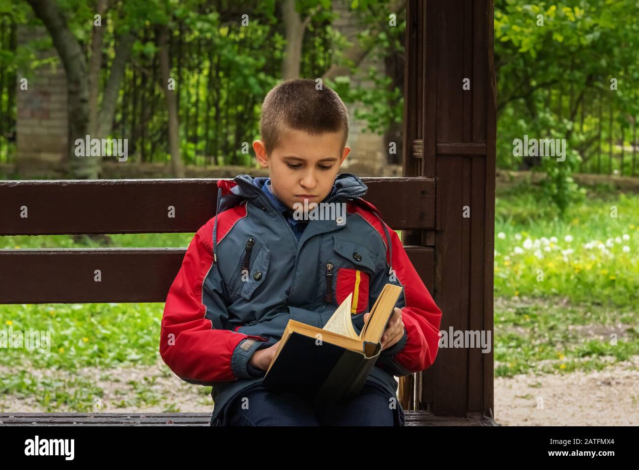 Ein ernsthafter Junge in legerer Kleidung liest gerade ein Buch. Der Junge sitzt allein auf einer alten, braunen Holzbank im Park. Selektiver Fokus. Unfokussiertes grünes Vol Stockfoto