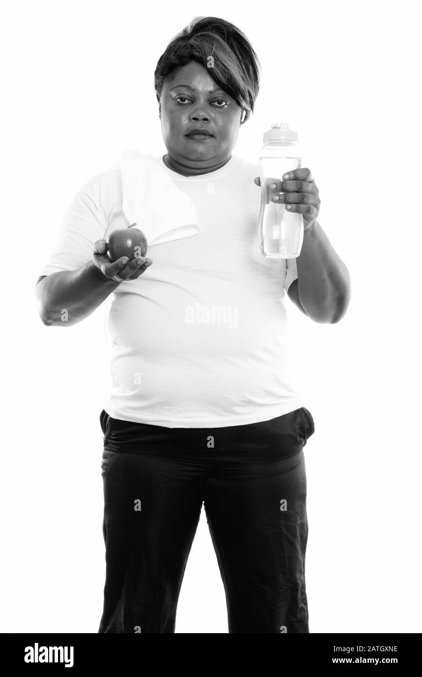 Studio-Schuss von fetter schwarzer Afrikanerin steht, während sie Wasserflasche und apfel für das Fitnessstudio hält Stockfoto