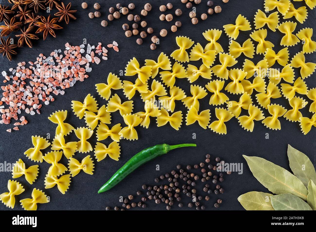 Stillleben mit roher Farfalle, verschiedenen Gewürzen und Salz auf einem schwarzen Steinfelder. Italienische, unbehackte Pasta. Lebensmittelkonzept oder Menüdesign. Flaches Lay. Stockfoto