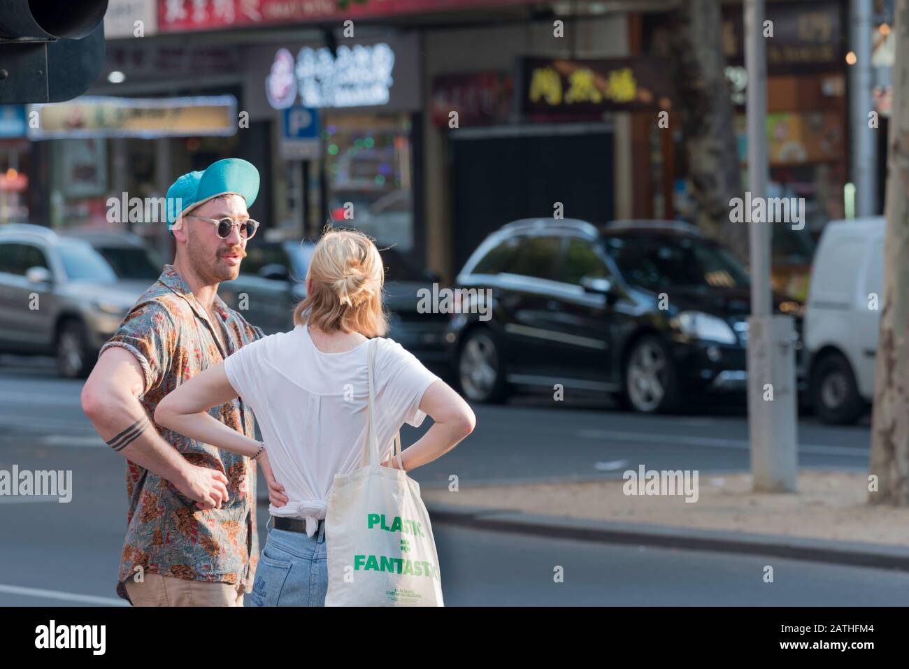 Ein junger Mann in Sonnenbrille und eine hochgedrehte Baseballmütze steht mit einer jungen Frau in einem weißen T-Shirt, an einer Straßenecke im inneren Melbourne, Australien Stockfoto