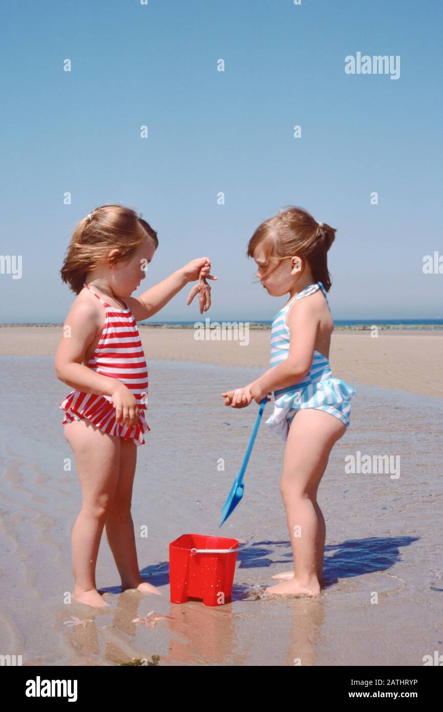 Kleine Mädchen, Zwillinge, in Badebekleidung an einem sandigen Strand, der einen Seestern hält und betrachtet Stockfoto