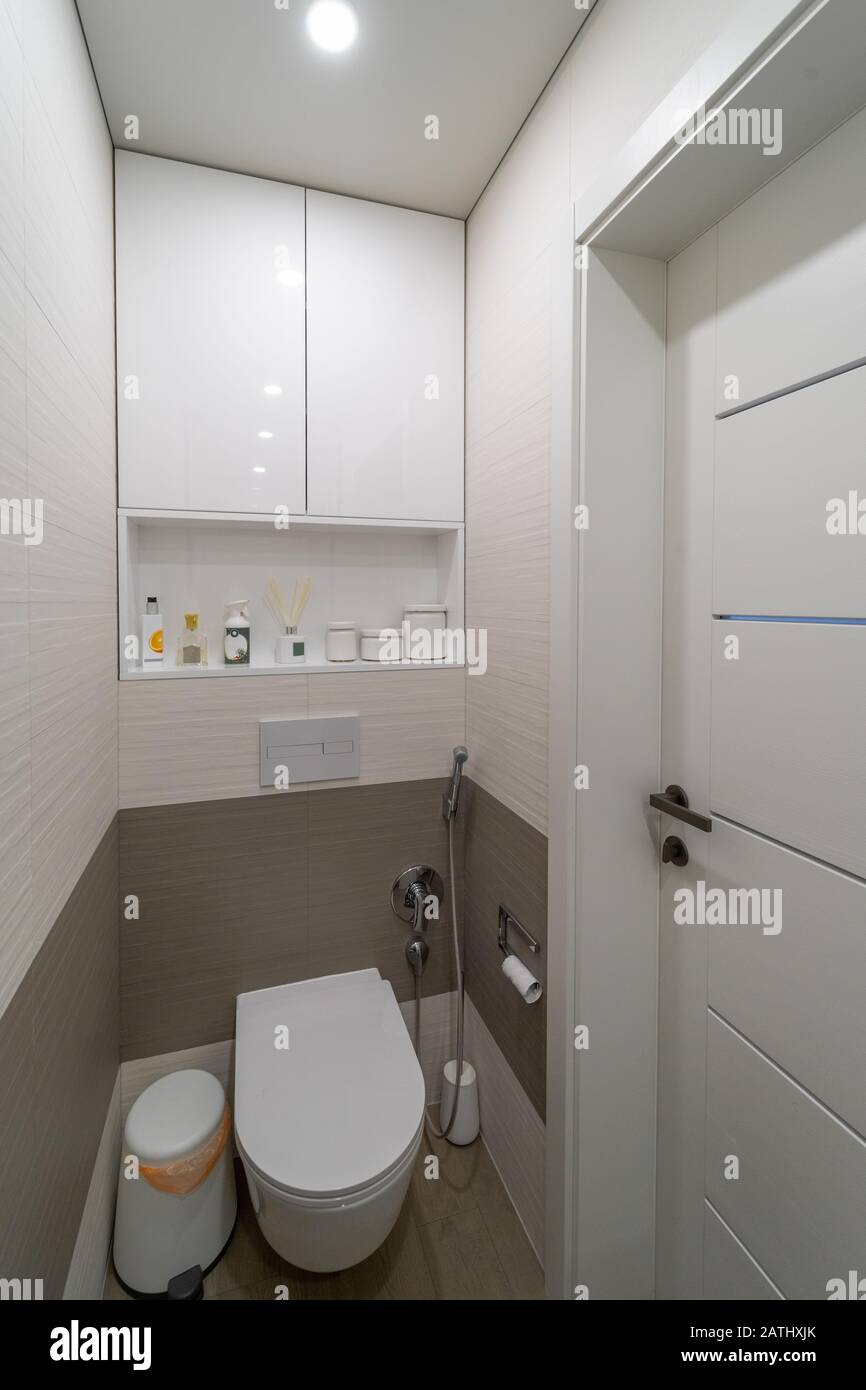 moderne designtoilette mit beigen und braunen Wand- und Bodenfliesen, Holztüren, weißer integrierter toilettenschüssel, bin, bidet Dusche, Schrubbbbürste und Lampe in Stockfoto