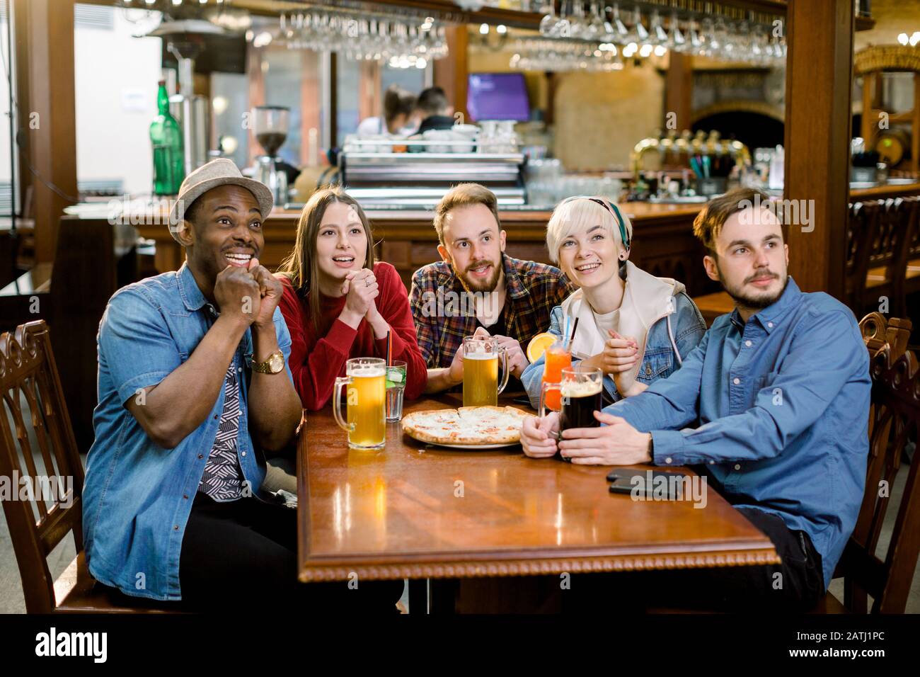 Fröhliche multirassische Freunde, die Spaß beim Essen in der Pizzeria haben. Fünf Freunde beobachten beim Essen von Pizza mit Getränken im gemütlichen Café gefühlvoll Sport im fernsehen Stockfoto