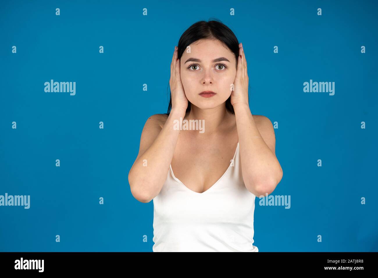 Vorderansicht der jungen Frau, die Ohren mit Händen bedeckt und die Kamera auf blauem Hintergrund isoliert betrachtet. Stockfoto