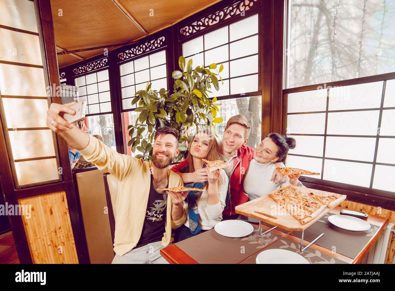 Die Firma der Jungs in der Pizzeria. Fröhliche Jungs mit Pizza in den Händen nehmen ein selfie auf ein Smartphone. Vier Freunde in einem Café Stockfoto