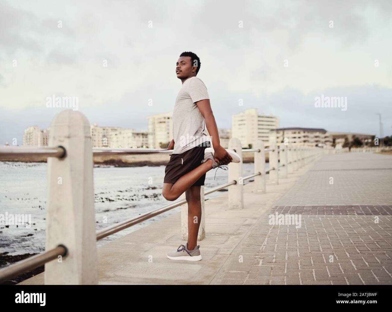Seitenansicht eines jungen männlichen Sportlers, der seine Beine streckt, der neben dem Geländer an der Strandpromenade steht Stockfoto