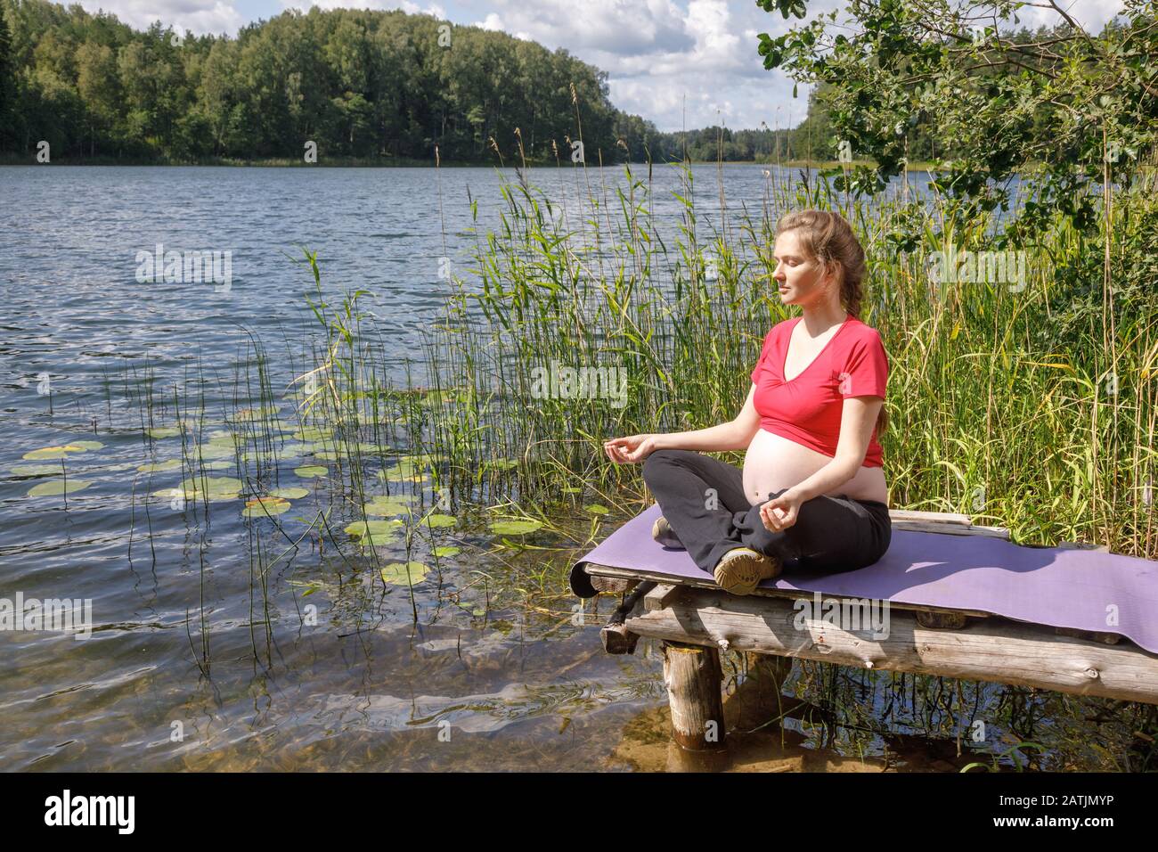 Junge schwangere Frau, die ein sportliches Kleid trägt und Yoga-Übungen macht, sitzt in lotus-position auf einem Holzsteg an einem Waldsee meditierend. Fitness und Heilung Stockfoto