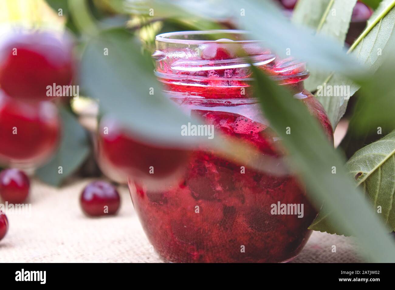 Kirschmarmelade in einem Glasbecher auf dem Hintergrund grüner Blätter auf einem Tisch aus Holztafeln. Nahaufnahme. Stockfoto