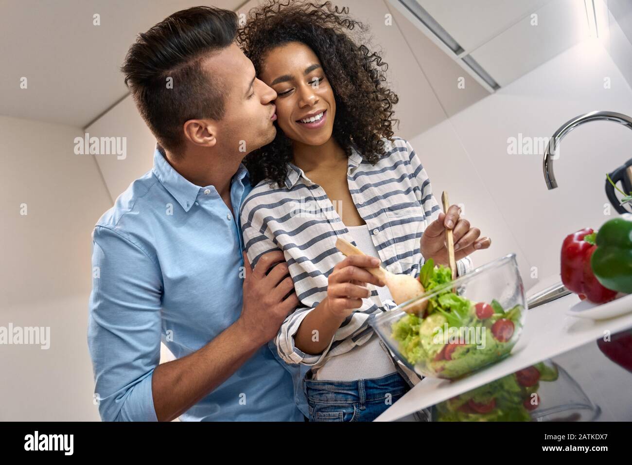 Junge Erwachsene mischten sich mit dem Paar, das küsst und gemeinsam gesunde Speisen kocht Stockfoto