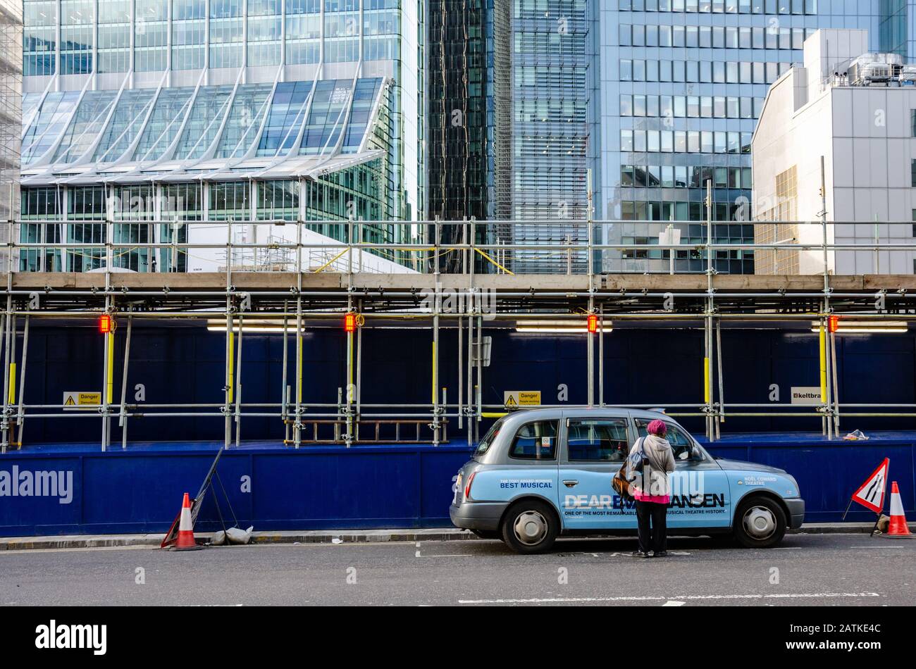 Eine Taxifahrerin steigt in ihr Taxi, das vor Bauarbeiten geparkt ist, und ist auf modernen Büroblöcken ausfahrend. Stockfoto