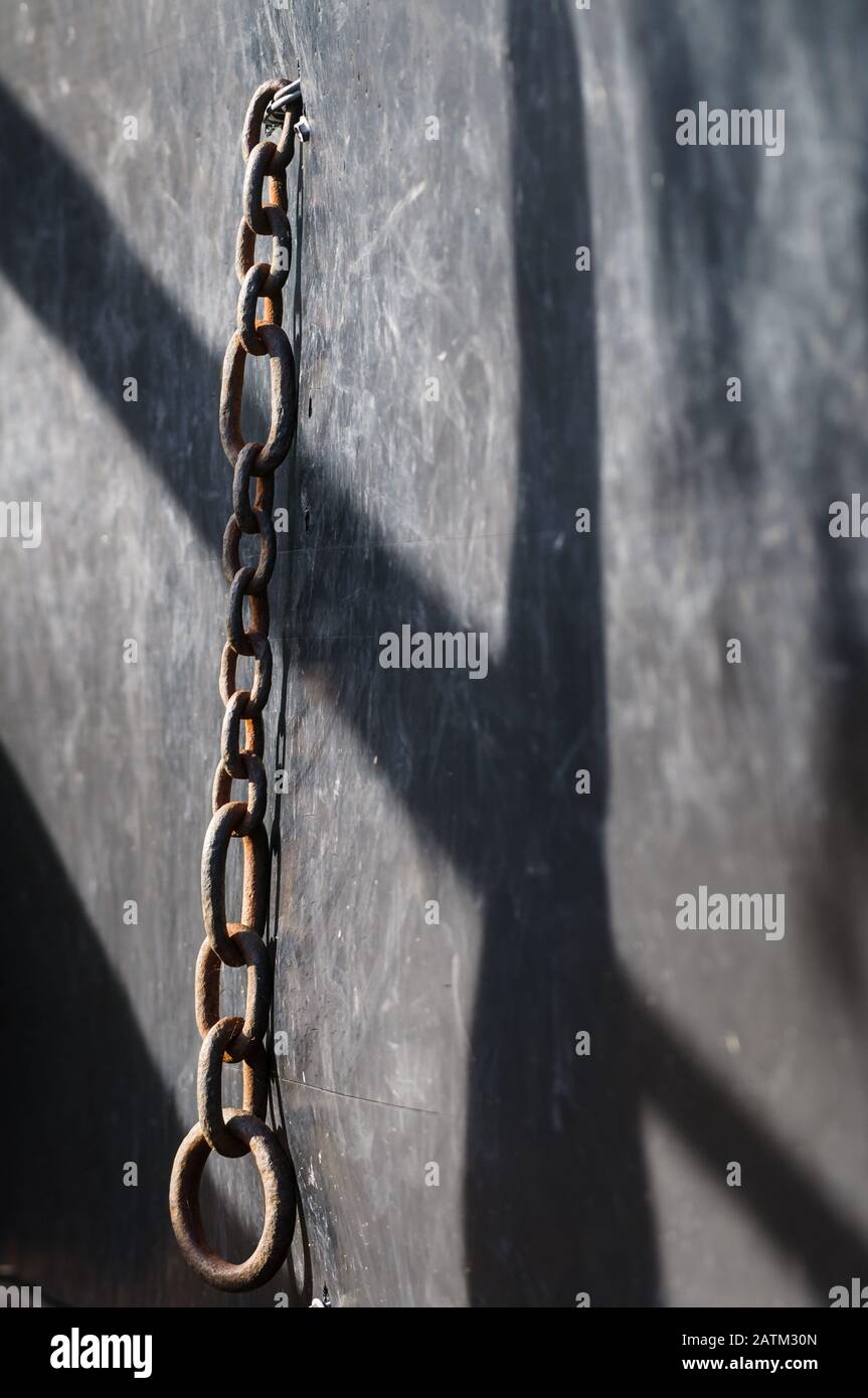 Einzelne Eisenkette hängt an einer überdachten Platte auf einem Viehhof mit dem ragenden Schatten der Schienen und Pfosten einer nahe gelegenen Platte der gleichen Viehhöfe. Stockfoto