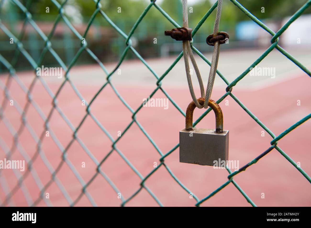 Nahaufnahme von verrostetem Vorhängeschloss am grünen Zaun mit Kabelanschluss. Tennisplatz im Hintergrund, Stockfoto