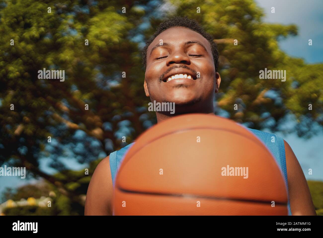 Porträt eines lächelnden, zufriedenen jungen männlichen Spielers mit geschlossenen Augen, der Basketball im Freien hält Stockfoto