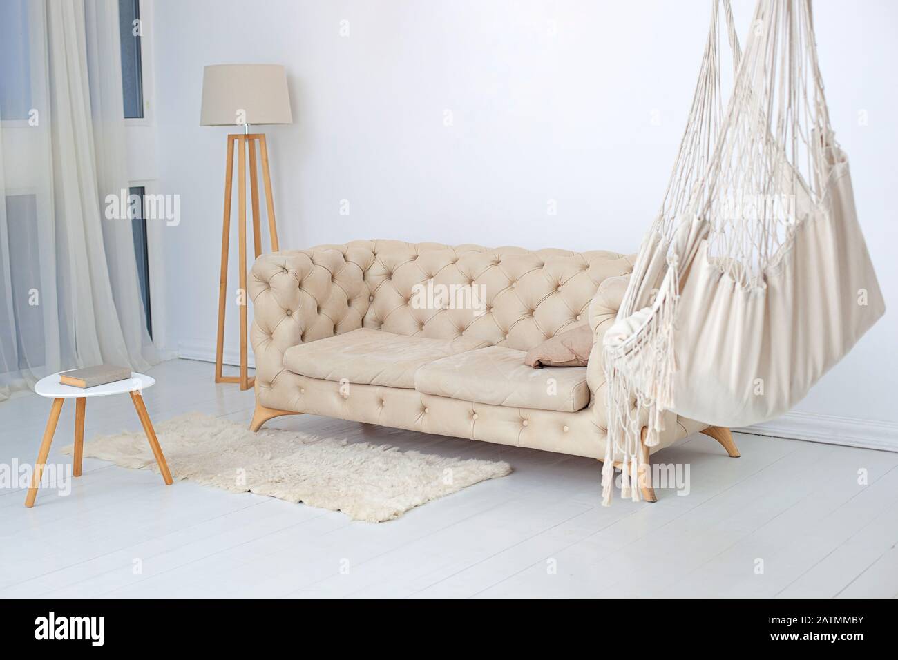 Wohnzimmer mit Hängematte, flauschiger Wolldecke und Couchtisch mit Lampe. Multifunktionales Loft-Apartment mit Hängematte, beigefarbenem Sofa und großem Fenster Stockfoto