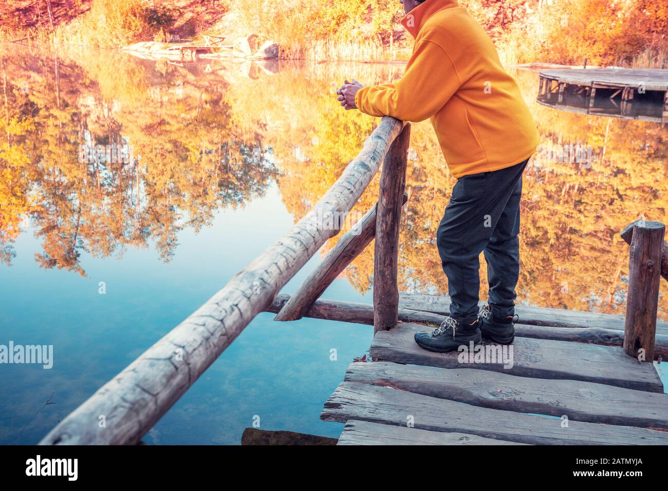 Ein Mann steht auf einem Holzdeck, das sich an ein Geländer lehnt und blickt auf einen Waldsee. Sonnenaufgang über dem See im Herbst Stockfoto