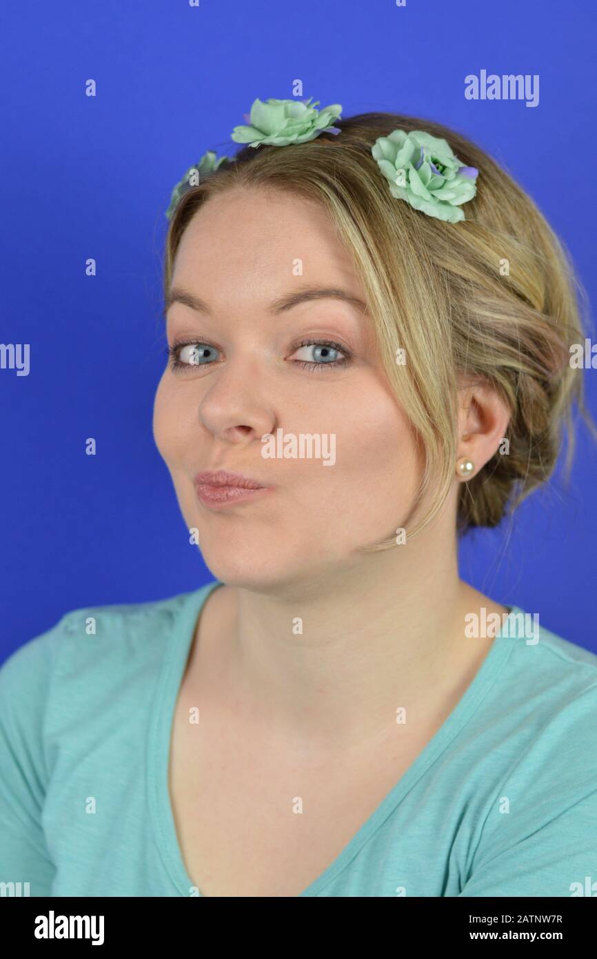 Porträt einer skeptischen jungen blonden kaukasischen Frau mit hochhaarigen Haaren und zyanblauen Blumen auf einem Haarzirklett, das vor blauem Hintergrund wiederkäut Stockfoto